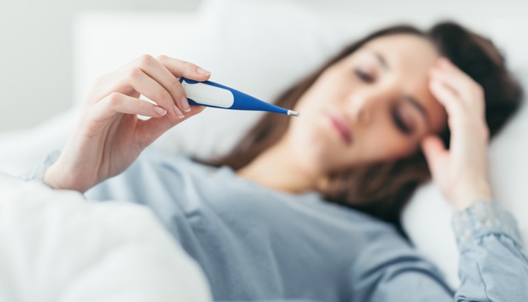 A woman who has the common cold is lying in bed looking at a fever thermometer